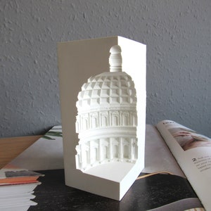 May include: White ceramic bookend shaped like a building with a dome. The bookend is designed to hold books upright.