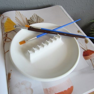 May include: White ceramic paint palette with a raised ridge for holding paintbrushes. Two paintbrushes are resting on the palette, one with a blue handle and one with a brown handle. The palette is on a white surface.