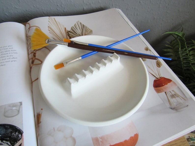 May include: White ceramic paint palette with a raised ridge for holding paintbrushes. Two paintbrushes are resting on the palette, one with a blue handle and one with a brown handle. The palette is on a white surface.