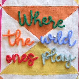 May include: A white knitted blanket with a colorful patchwork design. The words "Where the wild ones play" are spelled out in yarn across the blanket.