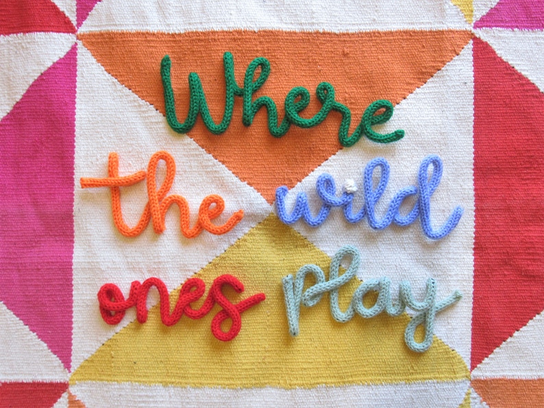 May include: A white knitted blanket with a colorful patchwork design. The words "Where the wild ones play" are spelled out in yarn across the blanket.