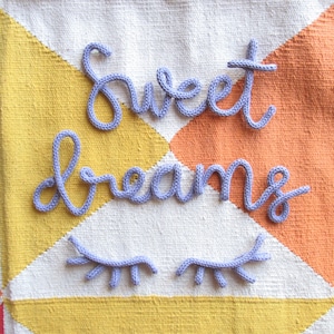 Könnte beinhalten: Gestrickter Text in Lavendelfarbe mit der Aufschrift "sweet dreams" und passenden gestrickten Wimpern. Der Text und die Wimpern sind auf einem farbenfrohen geometrischen Hintergrund mit gelben, orangefarbenen, weißen und roten Dreiecken platziert.