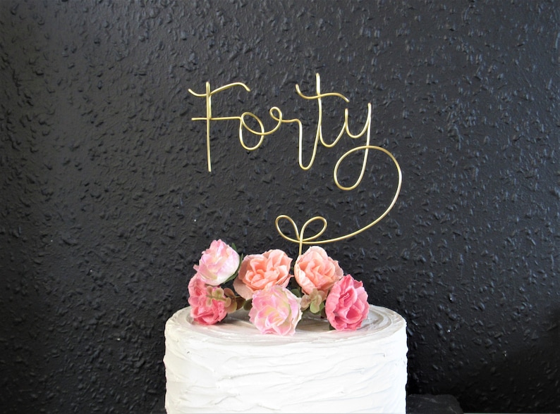 May include: Gold wire cake topper that says "Forty" with pink flowers on a white cake.