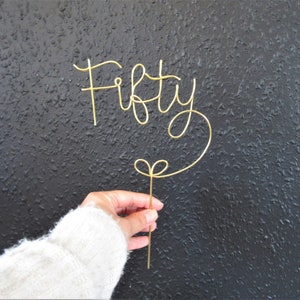 May include: Gold wire cake topper that says "Fifty" in a cursive font. The topper is attached to a thin wire that is bent into a heart shape at the bottom.