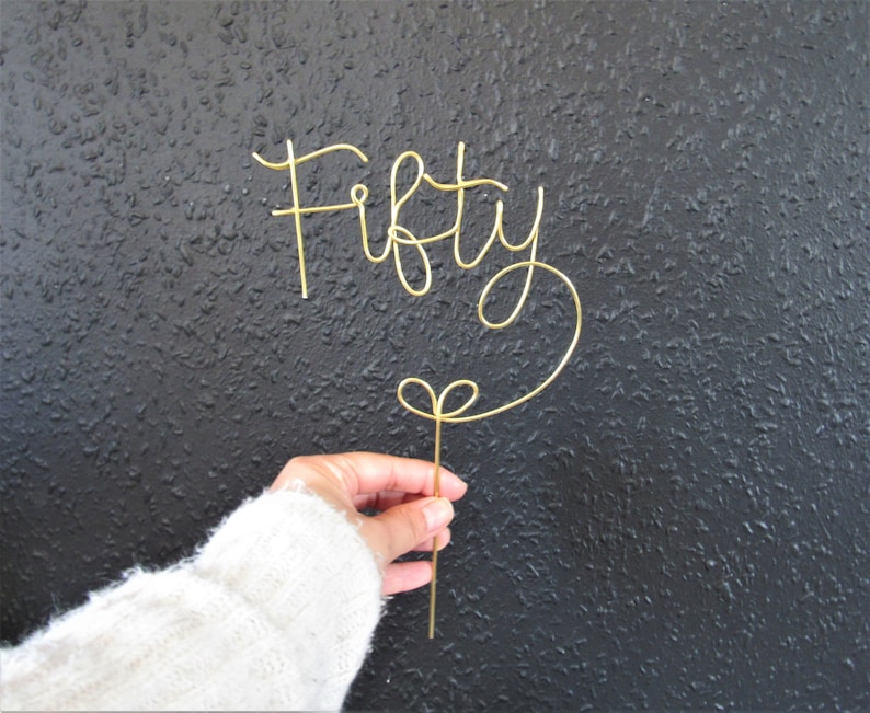 May include: Gold wire cake topper that says "Fifty" in a cursive font. The topper is attached to a thin wire that is bent into a heart shape at the bottom.