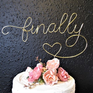 May include: Gold wire cake topper with the word "finally" and a heart. The topper is on a white cake with pink flowers.