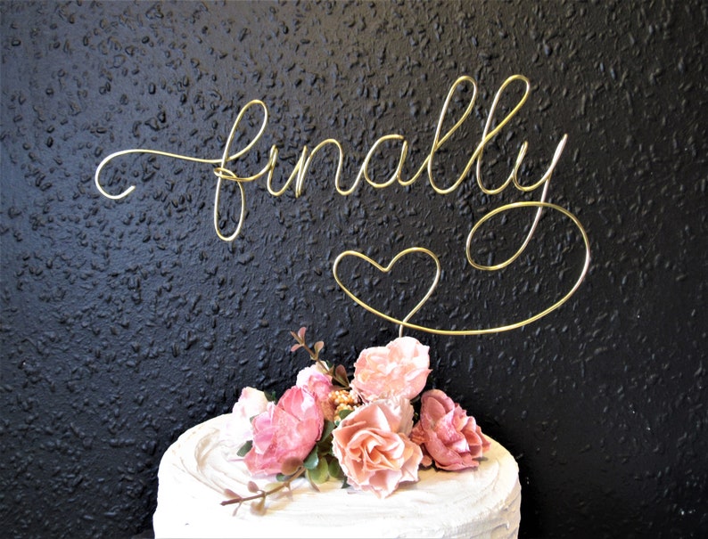 May include: Gold wire cake topper with the word "finally" and a heart. The topper is on a white cake with pink flowers.
