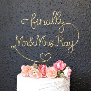 May include: Gold wire cake topper with the words "finally Mr & Mrs Ray" and a heart. The topper is on a white cake with pink flowers.
