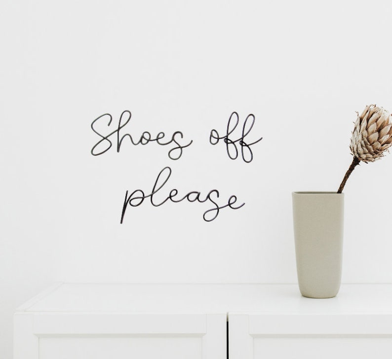 May include: White wall with black cursive lettering that reads "Shoes off please" and a beige vase with a dried flower arrangement.