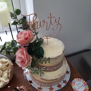 May include: A two-tiered white cake decorated with burlap ribbon and pink roses. A gold wire cake topper says "Thirty".