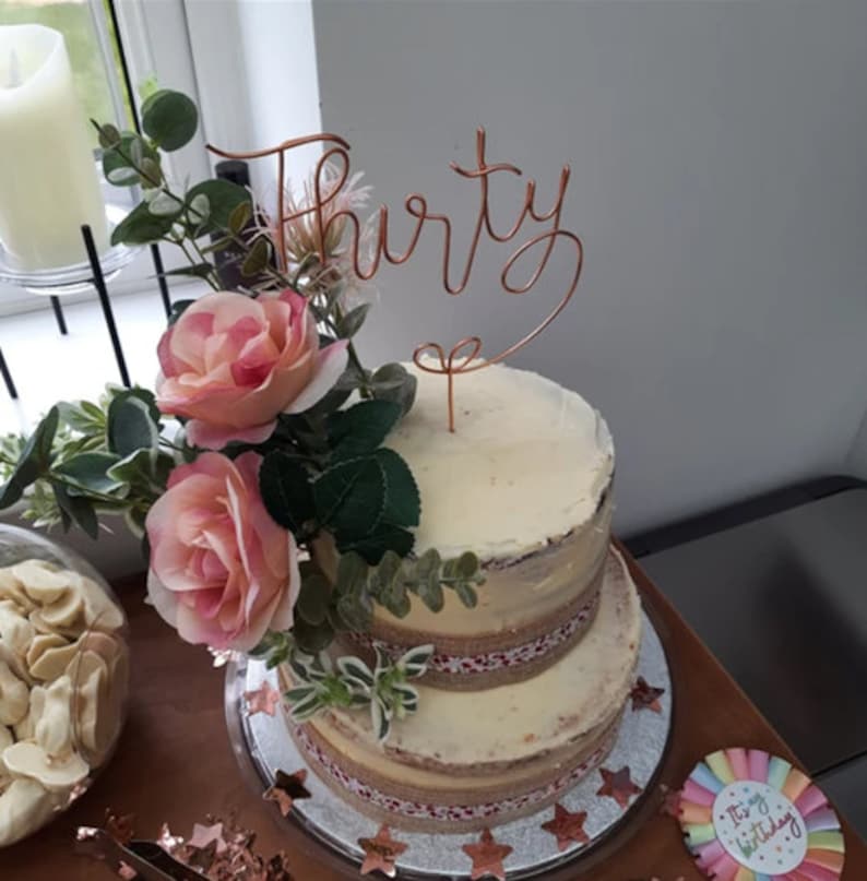May include: A two-tiered white cake decorated with burlap ribbon and pink roses. A gold wire cake topper says "Thirty".