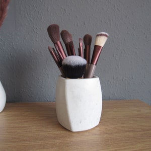 May include: A selection of makeup brushes with brown handles and white and black bristles are arranged in a white, geometric-shaped holder. The holder is on a wooden surface, with a grey wall behind.