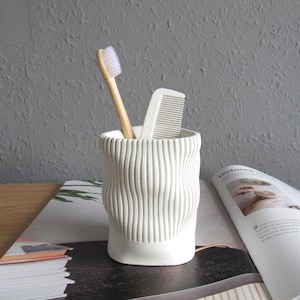 Może przedstawiać: Biały ceramiczny pojemnik na szczoteczkę do zębów z fakturowanym, falistym wzorem. W środku znajduje się biała szczoteczka do zębów i biały grzebień.