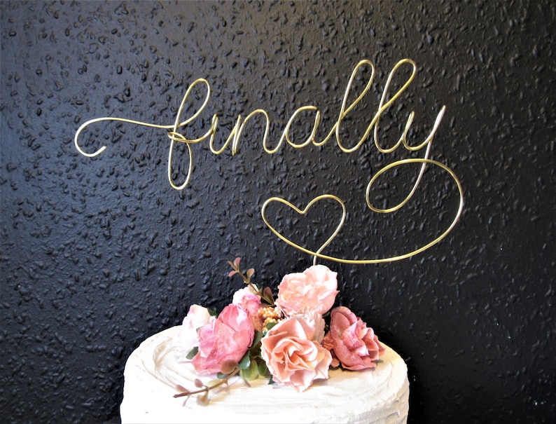 May include: Gold wire cake topper with the word "finally" and a heart. The topper is on a white cake with pink flowers.