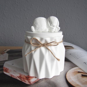 Könnte beinhalten: Weiße Keramikdose mit gefaltetem Design und einer kleinen weißen Hasenskulptur auf dem Deckel. Die Dose ist mit einem braunen Bindfaden gebunden.