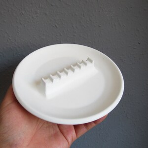 May include: White ceramic ashtray with a raised, rectangular, ridged design for holding cigarettes.