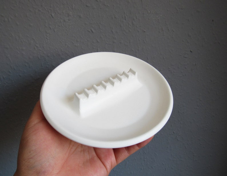 May include: White ceramic ashtray with a raised, rectangular, ridged design for holding cigarettes.