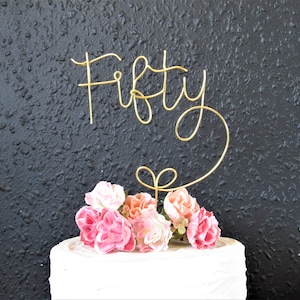 May include: Gold wire cake topper with the word "Fifty" in a cursive font. The topper is placed on a white cake decorated with pink and peach flowers.