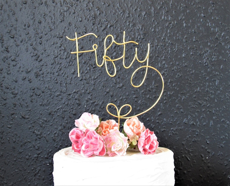 May include: Gold wire cake topper with the word "Fifty" in a cursive font. The topper is placed on a white cake decorated with pink and peach flowers.