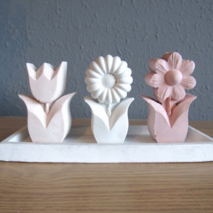 K&ouml;nnte beinhalten: Drei kleine, wei&szlig;e Keramik-Blumen-Skulpturen auf einem wei&szlig;en Tablett. Die Blumen sind eine rosa Tulpe, eine wei&szlig;e G&auml;nsebl&uuml;mchen und eine rosa Blume mit f&uuml;nf Bl&uuml;tenbl&auml;ttern.