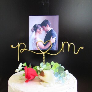 Könnte beinhalten: Ein Tortenaufsatz für eine Hochzeit mit einem Foto eines Paares, einem goldenen Drahtdesign mit den Buchstaben "p m" und einem Herz. Der Kuchen ist mit einer roten Rose, weißen Blumen und Grünzeug verziert. Der Hintergrund ist schwarz.
