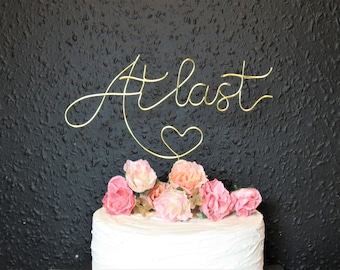 May include: A white cake with pink and peach flowers, topped with a gold wire phrase that reads "At last" with a heart. The cake sits against a textured black background.