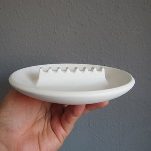 May include: White ceramic ashtray with a raised, wavy ridge in the center for holding cigarettes.