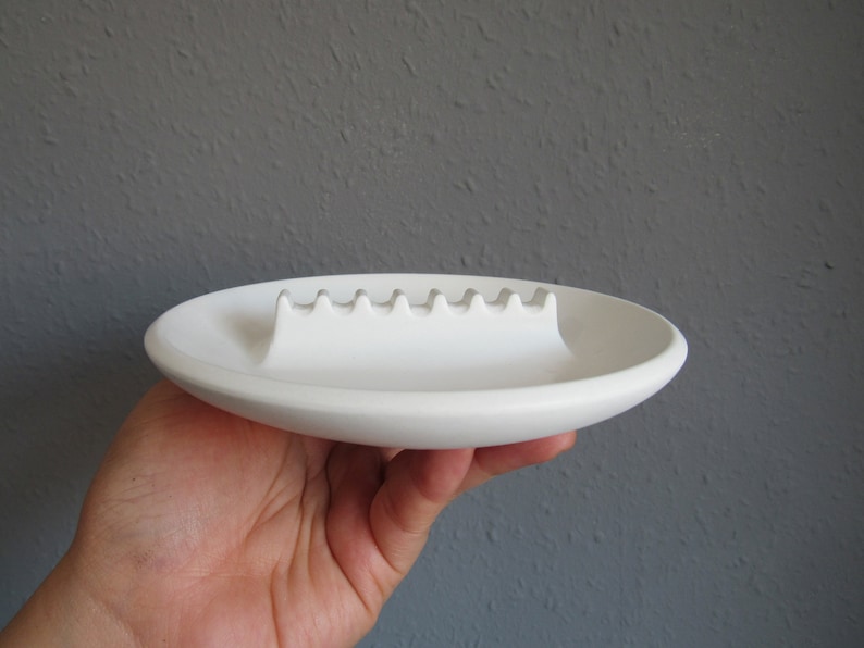 May include: White ceramic ashtray with a raised, wavy ridge in the center for holding cigarettes.