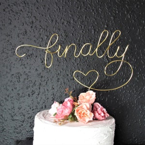 May include: Gold wire cake topper with the word "finally" and a heart. The topper is on a white cake with pink flowers.