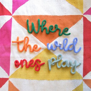 May include: A colorful knitted wall hanging with the words "Where the wild ones play" in a whimsical font.
