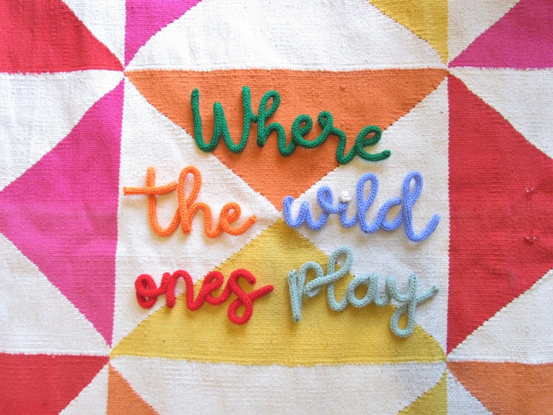 May include: A colorful knitted wall hanging with the words "Where the wild ones play" in a whimsical font.