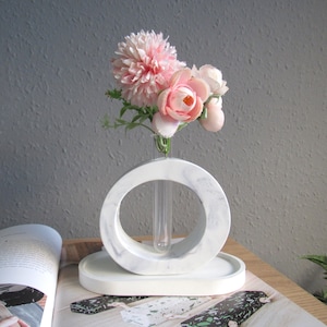 以下が含まれることがあります： 円形の開口部と中央にガラス製の試験管が付いた白いセラミック製のフラワーベース。ベースはマーブル模様で装飾されています。試験管にはピンクの花束が入っています。