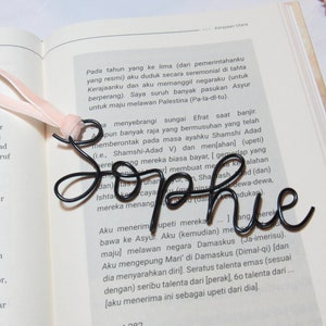 May include: A black metal bookmark shaped like the word "Sophie". The bookmark is resting on an open book with text in a foreign language.