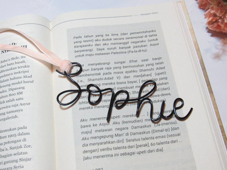 May include: A black metal bookmark shaped like the word "Sophie". The bookmark is resting on an open book with text in a foreign language.