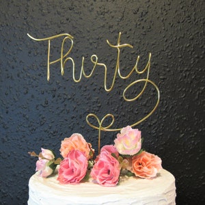 May include: Gold wire cake topper with the word "Thirty" in cursive. The topper is decorated with pink and peach artificial flowers.