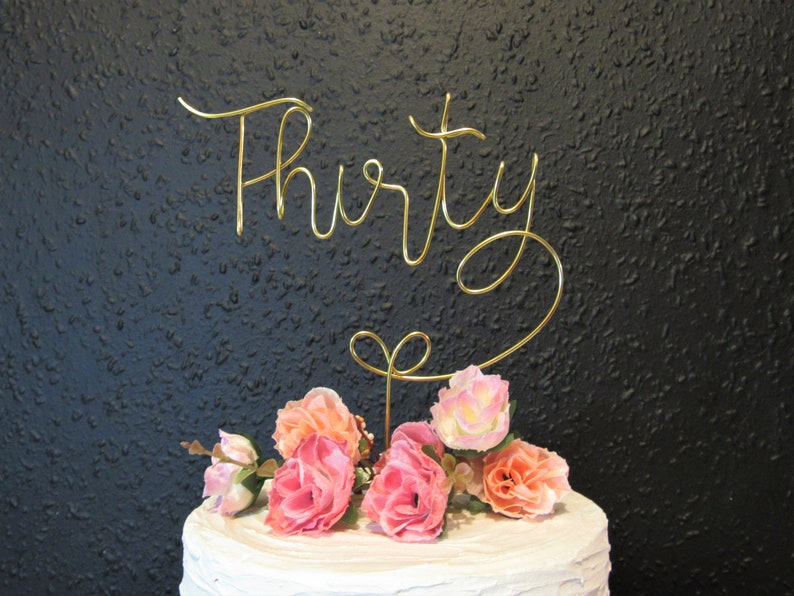 May include: Gold wire cake topper with the word "Thirty" in cursive. The topper is decorated with pink and peach artificial flowers.