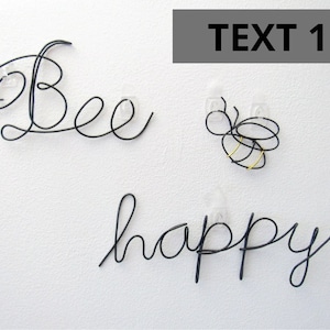 Puede incluir: Escultura de alambre negro de las palabras "Bee happy" con una escultura de abeja de alambre colgada arriba. La abeja tiene un cuerpo dorado.