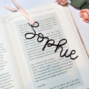 May include: A black metal bookmark shaped like the word "Sophie". The bookmark is laying on top of an open book with text in a foreign language.