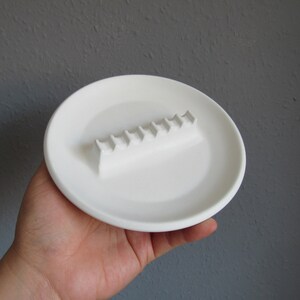 May include: White ceramic ashtray with a raised, rectangular section for holding cigarettes. The ashtray is about 6 inches in diameter.
