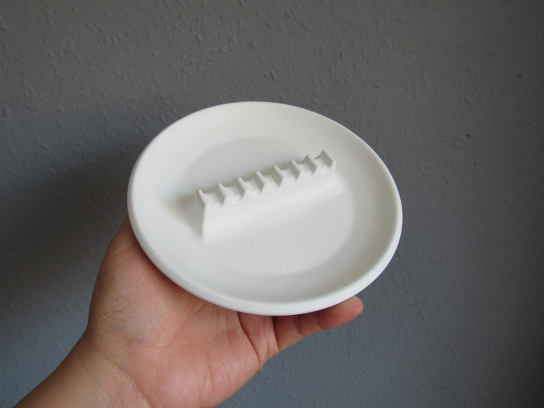 May include: White ceramic ashtray with a raised, rectangular section for holding cigarettes. The ashtray is about 6 inches in diameter.