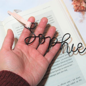 May include: A black wire bookmark shaped like the name "Sophie". The bookmark is held in a hand with a pink ribbon attached to the top.