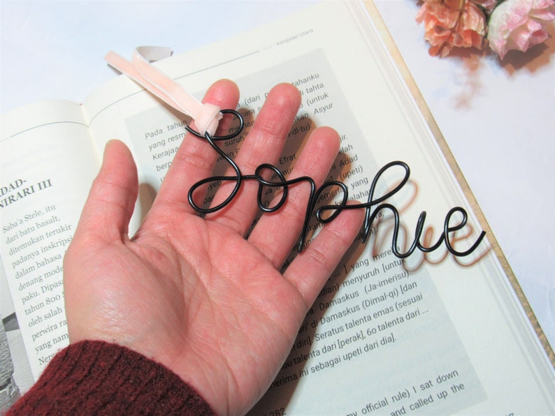 May include: A black wire bookmark shaped like the name "Sophie". The bookmark is held in a hand with a pink ribbon attached to the top.