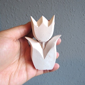 K&ouml;nnte beinhalten: Eine wei&szlig;e Tulpen-Skulptur aus Beton mit einem einfachen, geometrischen Design. Die Skulptur ist etwa 10 cm hoch und eignet sich perfekt, um Ihrem Zuhause einen Hauch von modernem Landhausstil zu verleihen.