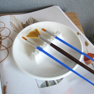 Puede incluir: Tres pinceles en un soporte de pinceles de cerámica blanca. Los pinceles tienen mangos azules y están dispuestos en un plato de cerámica blanca. El plato está sobre una superficie blanca.