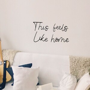 May include: A white couch with pillows and a guitar leaning against it. The wall behind the couch has the words "This feels like home" written on it in black.