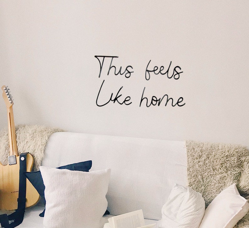 May include: A white couch with pillows and a guitar leaning against it. The wall behind the couch has the words "This feels like home" written on it in black.