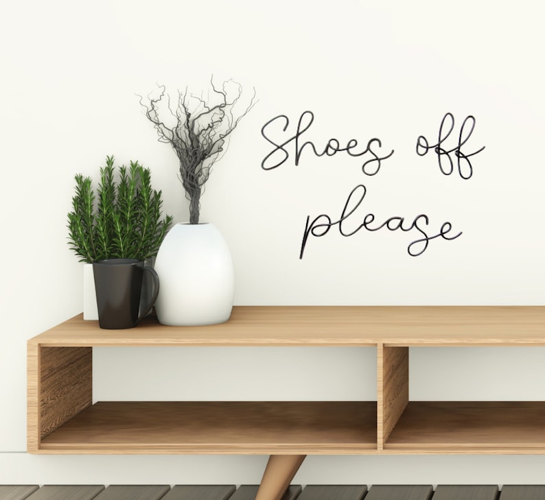 May include: A wooden shelf with two compartments, a white vase with dried branches, a black mug, and a green plant. The wall behind the shelf has a black cursive sign that reads "Shoes off please".