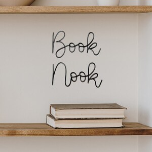 Puede incluir: Una estanter&iacute;a de madera con una pared blanca detr&aacute;s. La estanter&iacute;a tiene un letrero que dice "Book Nook" en letras cursivas negras. Hay tres libros apilados en la estanter&iacute;a.