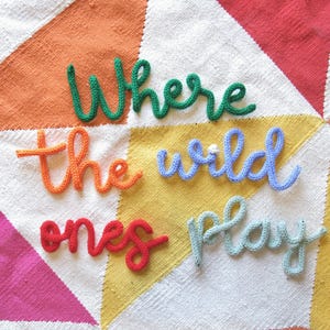 May include: A white knitted blanket with a colorful geometric pattern. The words "Where the wild ones play" are spelled out in yarn on the blanket.