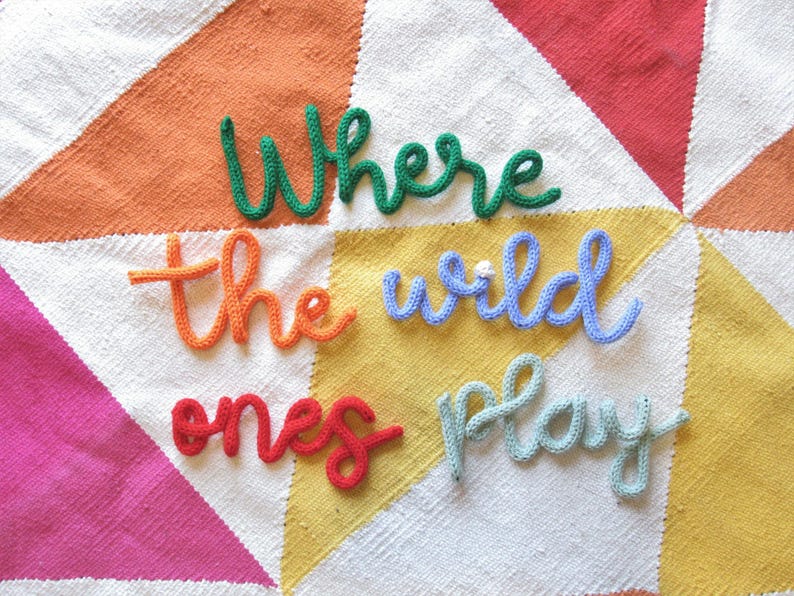 May include: A white knitted blanket with a colorful geometric pattern. The words "Where the wild ones play" are spelled out in yarn on the blanket.